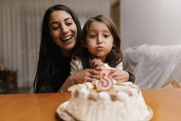 Girl blowing candle over birthday cake while sitting with mother at home - EGAF01525