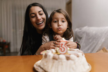 Mädchen bläst eine Kerze auf einem Geburtstagskuchen aus, während sie mit ihrer Mutter zu Hause sitzt - EGAF01525