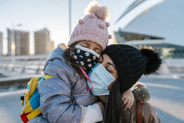 Mutter und Tochter mit Strickmütze und Gesichtsmaske umarmen sich, während sie im Freien stehen - EGAF01527