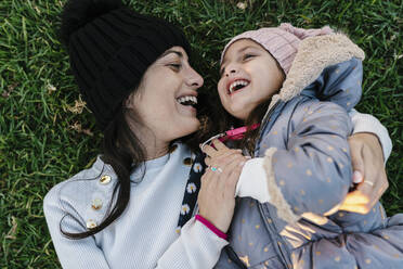 Smiling mother wearing knit hat playing with daughter while lying on grass - EGAF01528