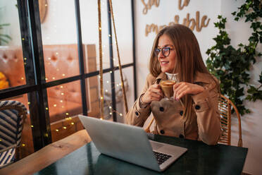 Lächelnde junge Bloggerin in Freizeitkleidung und mit Brille sitzt am Tisch mit einer Tasse Kaffee und tippt auf der Laptop-Tastatur, während sie Inhalte für soziale Netzwerke in einer gemütlichen Cafeteria vorbereitet - ADSF20358