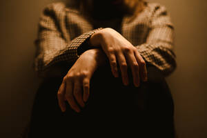 Selektiver Fokus der Hände einer anonymen, erschöpften, depressiven, unglücklichen Frau in einem karierten Mantel, die in einem dunklen Studio sitzt - ADSF20363