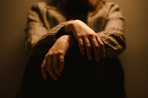 Selective focus of hands of crop anonymous exhausted depressive unhappy woman in checkered coat sitting in dark studio - ADSF20363