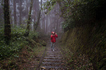 Full body of Asian female in raincoat climbing stairway and exploring nature while hiking through green forest in rainy day - ADSF20374