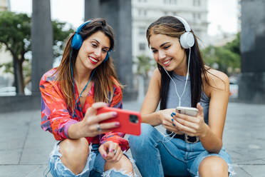 Fröhliche Hipster-Studentinnen in trendigen Outfits, die in sozialen Netzwerken auf ihren Handys surfen und mit Kopfhörern Musik hören, während sie im Sommer auf einer städtischen Straße zusammensitzen - ADSF20382