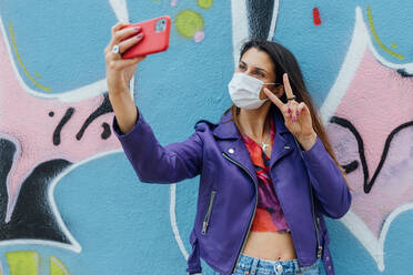 Trendige Millennial-Frau in modischer Kleidung und mit medizinischer Einwegmaske, die in der Nähe einer bunt bemalten Graffiti-Wand steht und ein Selfie mit ihrem Smartphone macht - ADSF20383