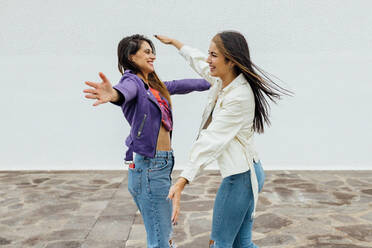 Cheerful young female friends in trendy outfits embracing each other and laughing while meeting on urban street near white stone wall looking at each other - ADSF20384