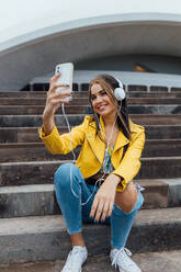 Fröhliche Millennial-Frau in trendigen bunten Outfits und Kopfhörer sitzen auf Steintreppen und nehmen Selfie auf Smartphone, während die Zeit zusammen in der Stadt - ADSF20386