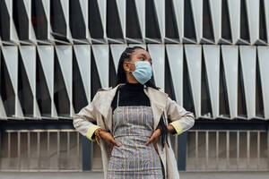 Low angle of confident young African American female in casual outfit and disposable protective mask looking away while standing against wall of modern urban building - ADSF20391