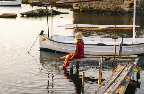 Woman wearing hat looking at view while sitting on pier - DGOF01837