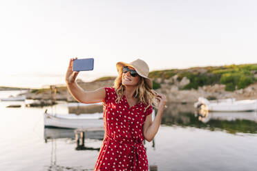 Lächelnde Frau mit Hut, die ein Selfie mit ihrem Mobiltelefon macht, während sie gegen den Himmel steht - DGOF01844