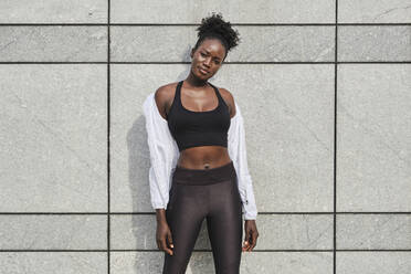 Eine fitte afroamerikanische Sportlerin steht auf einer Betonmauer auf der Straße, während sie sich nach dem Training ausruht und in die Kamera schaut - ADSF20399