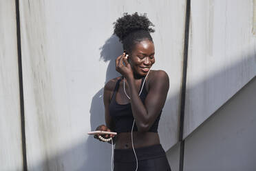 Lächelnde, selbstbewusste schwarze Sportlerin mit Kopfhörern, die Lieder auf dem Handy hört, während sie an einem sonnigen Tag auf der Straße steht - ADSF20401