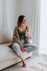 Smiling tender female in domestic outfit resting on couch with cup of hot drink while enjoying morning at home and looking away - ADSF20420