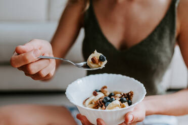 Abgeschnittene, nicht erkennbare Frau mit einer leckeren Schüssel mit gesundem Müsli, die zu Hause auf dem Boden sitzt und ein nahrhaftes Frühstück zu sich nimmt - ADSF20422