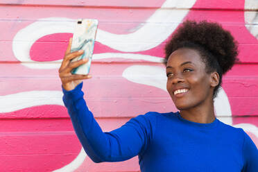 Confident African American female millennial taking self portrait on smartphone camera while standing near graffiti wall in urban area - ADSF20450