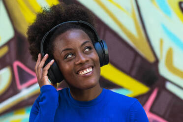 Optimistische schwarze Frau mit Afrofrisur, die mit Kopfhörern Musik hört, während sie auf einem unscharfen Hintergrund einer Graffitiwand auf der Straße steht - ADSF20451