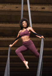 Slender African American female balancing on hanging hammock while doing aerial yoga above ground under bridge in city - ADSF20452