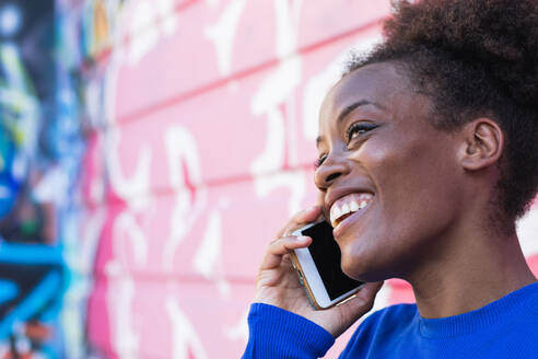 Sorglose afroamerikanische Frau mit Afrofrisur, die mit einem Freund über Nachrichten auf dem Smartphone diskutiert, während sie auf der Straße in der Nähe eines Gebäudes mit Graffiti steht und wegschaut - ADSF20454