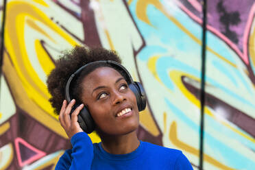 Optimistische schwarze Frau mit Afrofrisur, die mit Kopfhörern Musik hört, während sie auf einem unscharfen Hintergrund einer Graffitiwand auf der Straße steht - ADSF20455