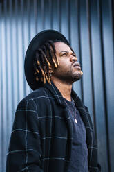 Informal bearded ethnic man with dreadlocks and in hat looking away standing on street - ADSF20458