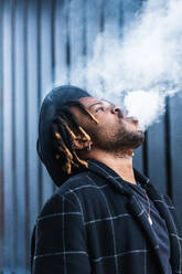 Seitenansicht eines ethnischen Mannes mit Dreadlocks und Hut in einer dichten Rauchwolke, der auf einer Straße in der Stadt raucht - ADSF20459