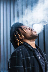 Side view of ethnic back guy with dreadlocks and hat in dense cloud of fume smoking on street in city - ADSF20459