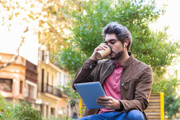 Ruhiger Mann in legerem Stil, der in der Stadt sitzt und einen Kaffee zum Mitnehmen in einem Pappbecher genießt, während er auf einem Tablet surft - ADSF20463