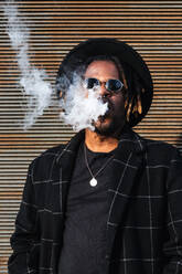 Modern African American man with dreadlocks wearing hat with sunglasses and exhaling smoke on street - ADSF20466