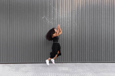 Joyful African American female with curly hair scattering white confetti in city while having fun and looking away - ADSF20469