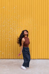 Joyful African American female with curly hair scattering white confetti in city while having fun and looking up - ADSF20477