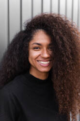 Charming African American female in black trendy outfit and with curly hair standing on street and looking at camera - ADSF20482