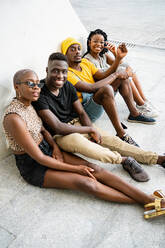 Gruppe afroamerikanischer Freunde in cooler, stilvoller Kleidung, die in der Nähe eines städtischen Gebäudes in der Stadt sitzen, während sie sich am Wochenende treffen und gemeinsam chillen und in die Kamera schauen - ADSF20491