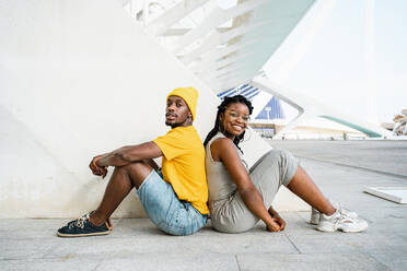 Side view of cool African American male and female friends sitting back to back on the street and looking at camera - ADSF20493