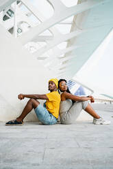 Side view of cool African American male and female friends sitting back to back on the street and looking at camera - ADSF20494