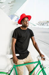 Confident African American male in trendy clothes standing with modern bike in urban area and looking away - ADSF20507