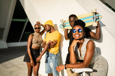 Group of African American friendly people in stylish summer clothes standing together on street with longboard and modern bike while looking at camera - ADSF20509