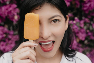 Happy woman with mouth open covering eye with orange ice cream - DSIF00269