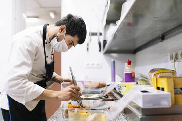 Männlicher Koch bei der Zubereitung von Speisen in Plastikbehältern in der Küche eines Restaurants während COVID-19 - DGOF01865