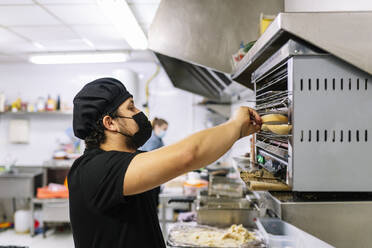 Männlicher Koch bei der Zubereitung von Brot im Ofen in der Restaurantküche während COVID-19 - DGOF01883