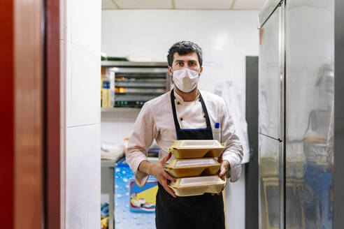 Junger Koch in Schürze mit Lebensmittelbehältern bei der Arbeit in einem Restaurant während einer Pandemie - DGOF01887