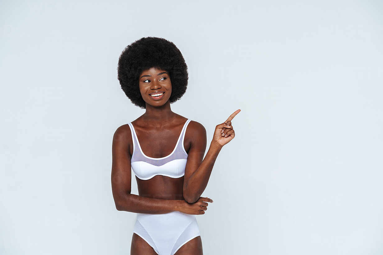 Skinny woman with afro hair in lingerie pointing while standing against  white background stock photo