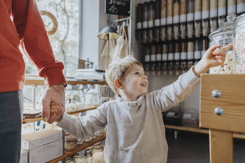 Fröhlicher Junge, der die Hand seines Vaters hält, während er auf ein Glas mit Süßigkeiten in einem Geschäft zeigt - MFF06900