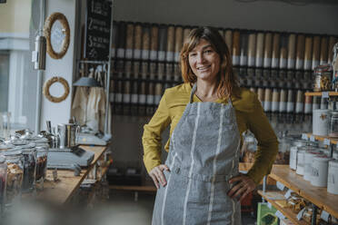 Smiling female entrepreneur in apron with hand on hip standing in zero waste shop - MFF06945