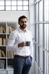 Lächelnder männlicher Unternehmer mit Händen in den Taschen, der eine Kaffeetasse im Büro hält - GIOF10834
