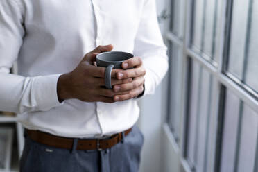 Junger Geschäftsmann, der eine Kaffeetasse hält, während er am Arbeitsplatz steht - GIOF10835