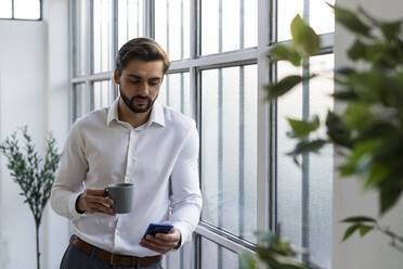 Männlicher Unternehmer mit Kaffeetasse, der sein Smartphone benutzt, während er sich bei der Arbeit an ein Fenster lehnt - GIOF10837