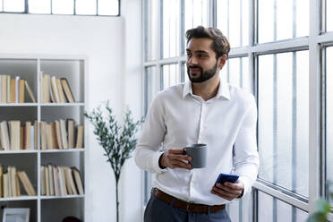 Lächelnder Geschäftsmann mit Mobiltelefon, der eine Kaffeetasse hält, während er im Büro wegschaut - GIOF10838