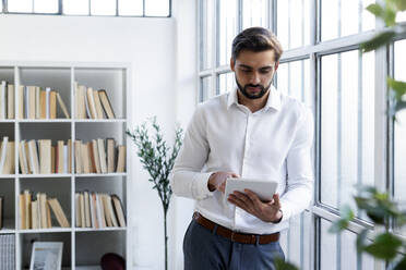 Männlicher Berufstätiger, der an einem digitalen Tablet arbeitet, während er sich bei der Arbeit an ein Fenster lehnt - GIOF10839