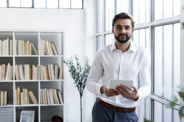 Smiling male entrepreneur holding digital tablet while leaning on glass window at work place - GIOF10840